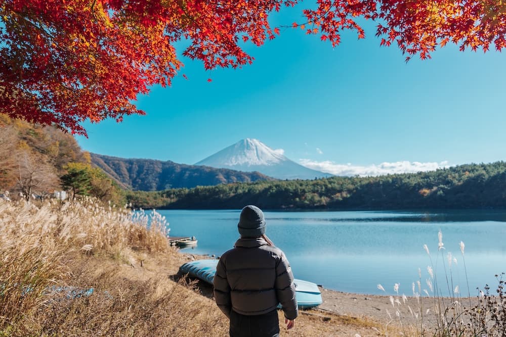 Frau-mit-Mutze-und-Daunenjacke-blickt-uber-einen-See-auf-den-schneebedeckten-Fuji,-umrahmt-von-roten-Ahornblattern-im-Herbst.jpg